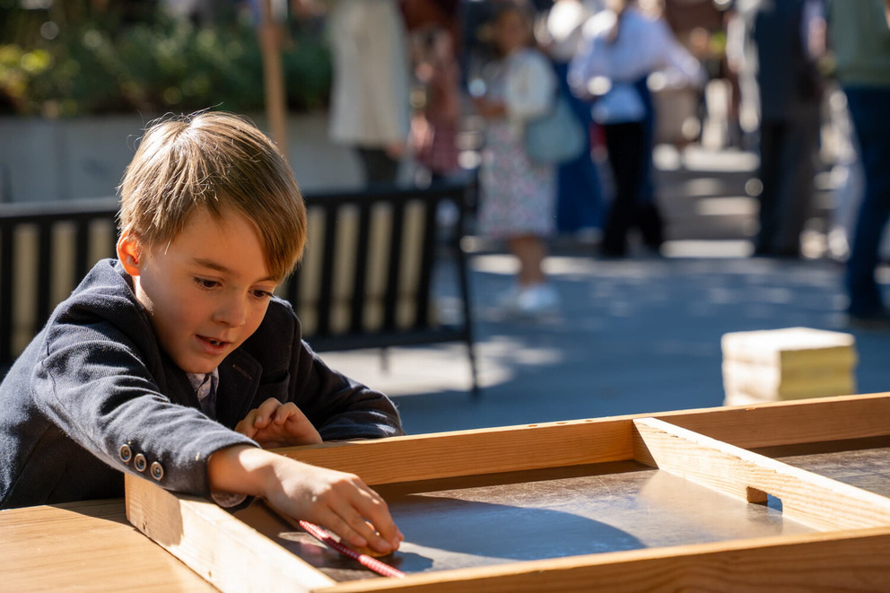 Een jongen speelt geconcentreerd met een houten spel op een buitenlocatie, in een drukke omgeving.