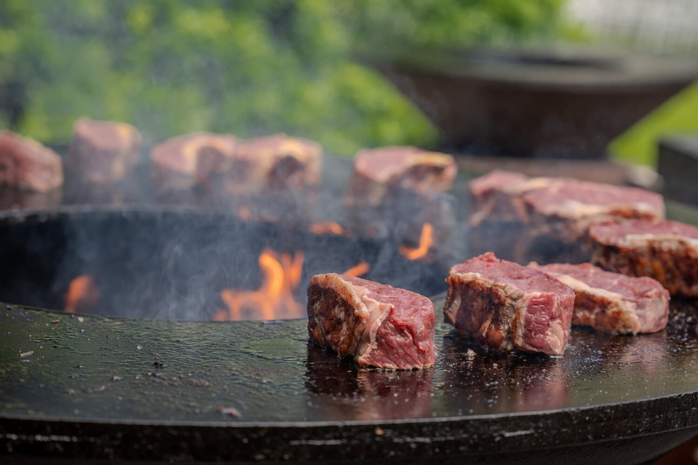 Stukken vlees grillen op een roestvrijstalen barbecue, omringd door rook en vlammen.