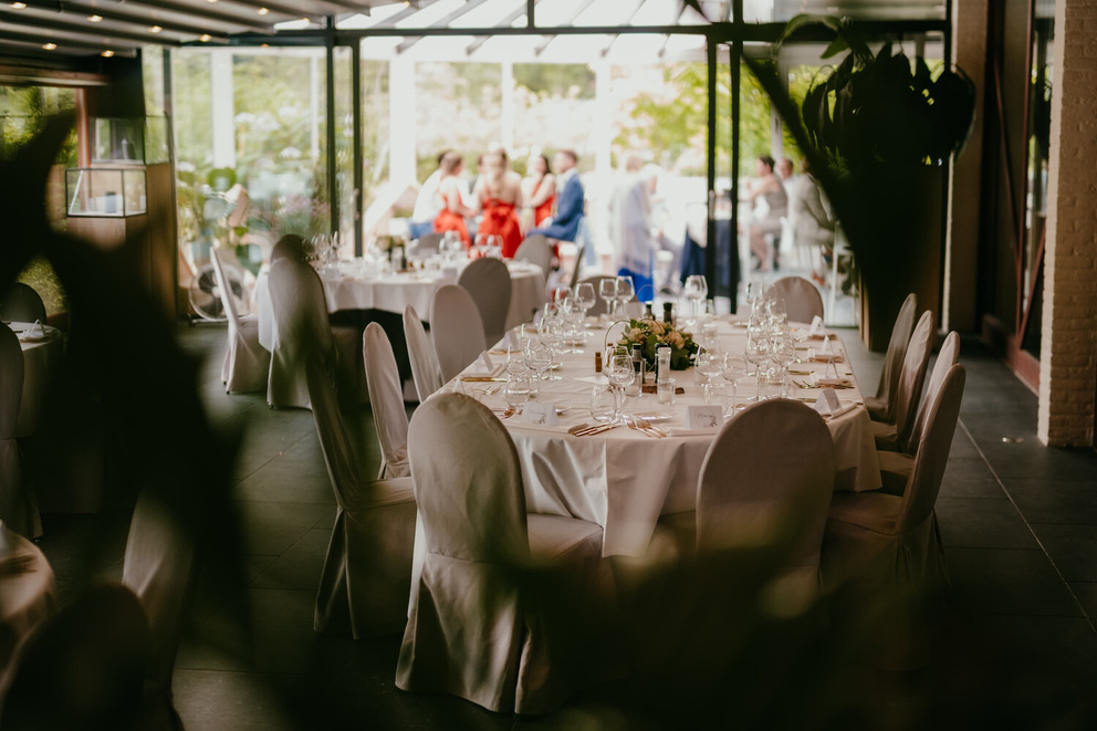 Stijlvol gedekte eettafels in een elegante ruimte met gasten op de achtergrond. Groene planten op de voorgrond.