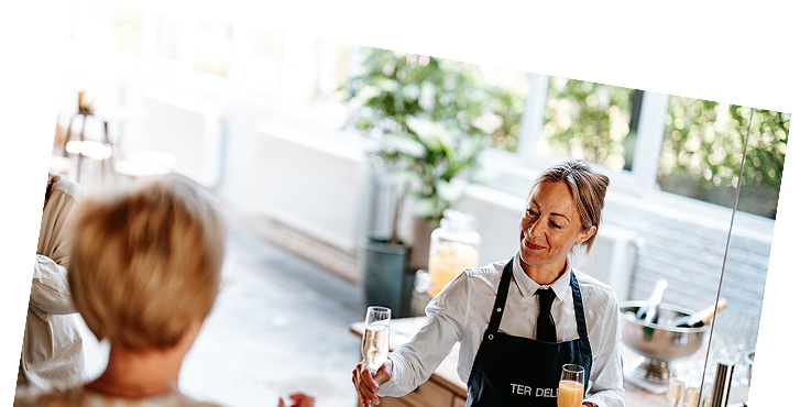 Een serveerster met een schort biedt een drankje aan aan een klant in een lichte, moderne caféomgeving.
