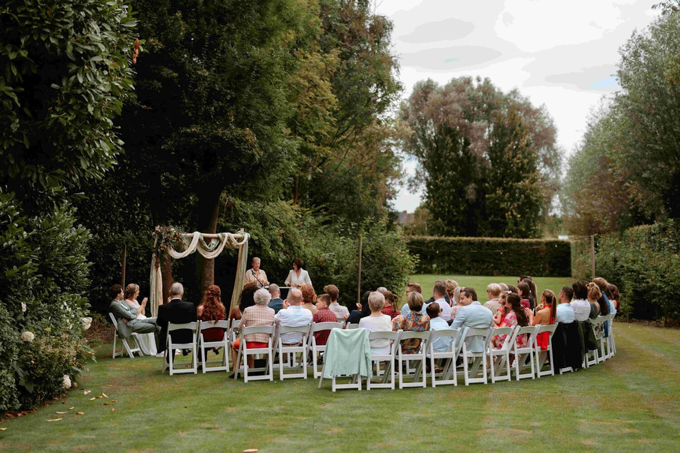 Buitenceremonie met gasten in witte stoelen, omringd door groen, terwijl een stel hun geloften uitspreekt.