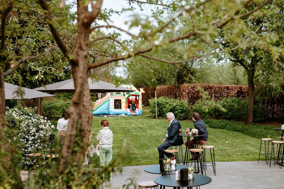 Twee mensen zitten op hoge stoelen terwijl anderen kijken naar een springkasteel in een groene tuin.
