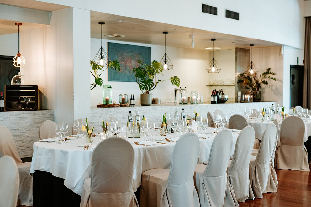 Elegant ingericht restaurant met een lange eettafel, witte tafelkleden en moderne verlichting.