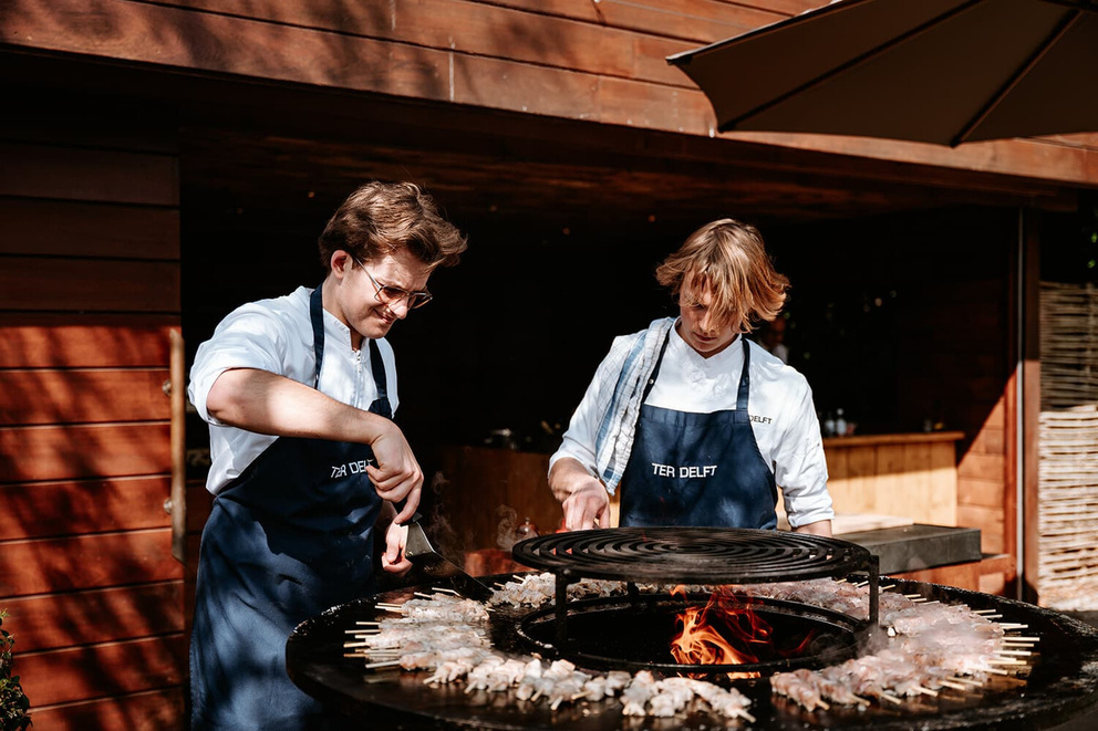Twee mannen in witte overhemden en schorten bereiden voedsel op een grote grill buiten.