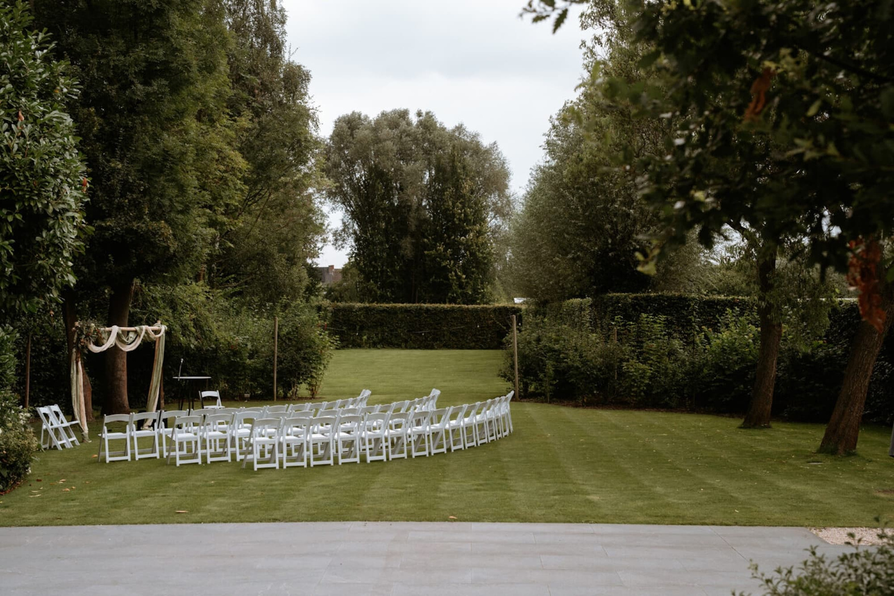 Een groene tuin met een cirkelvormige opstelling van witte stoelen en een decoratieve boog voor een ceremonie.