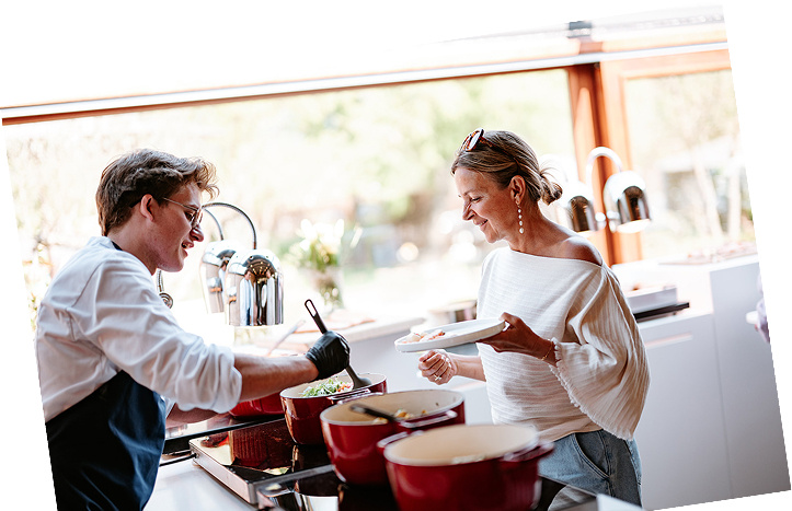 Een jonge man kookt in een moderne keuken terwijl een vrouw hem glimlachend aankijkt en een bord vasthoudt.