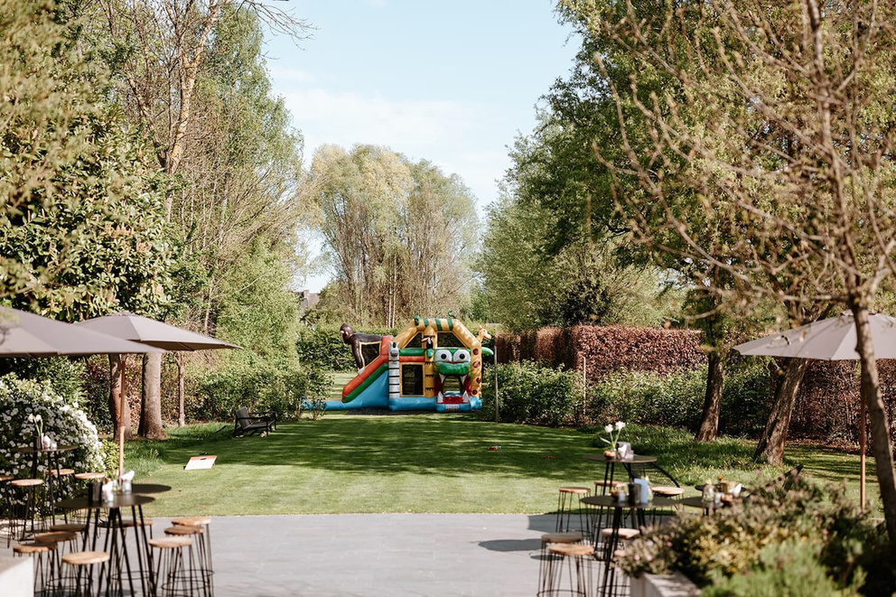 Landschap met een groene tuin, een opblaasbare glijbaan en enkele tafels onder parasols op de voorgrond.