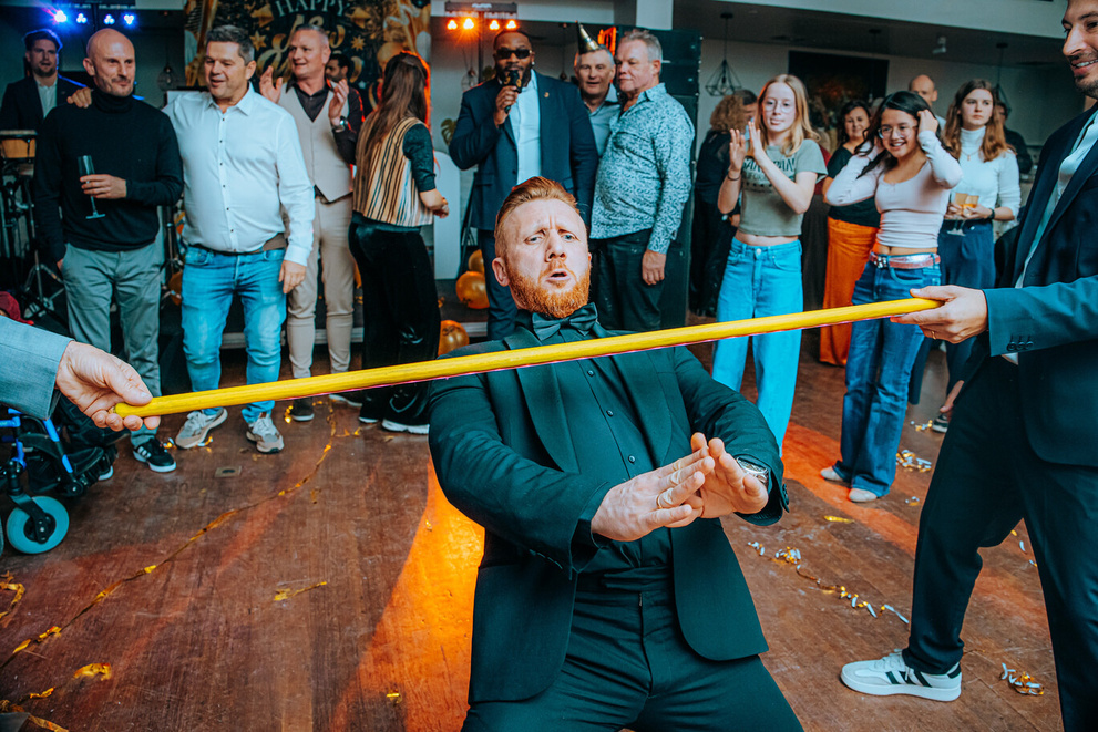 Een man in een zwart pak doet een limbo-dans onder een gele stok, omringd door een feestende menigte.
