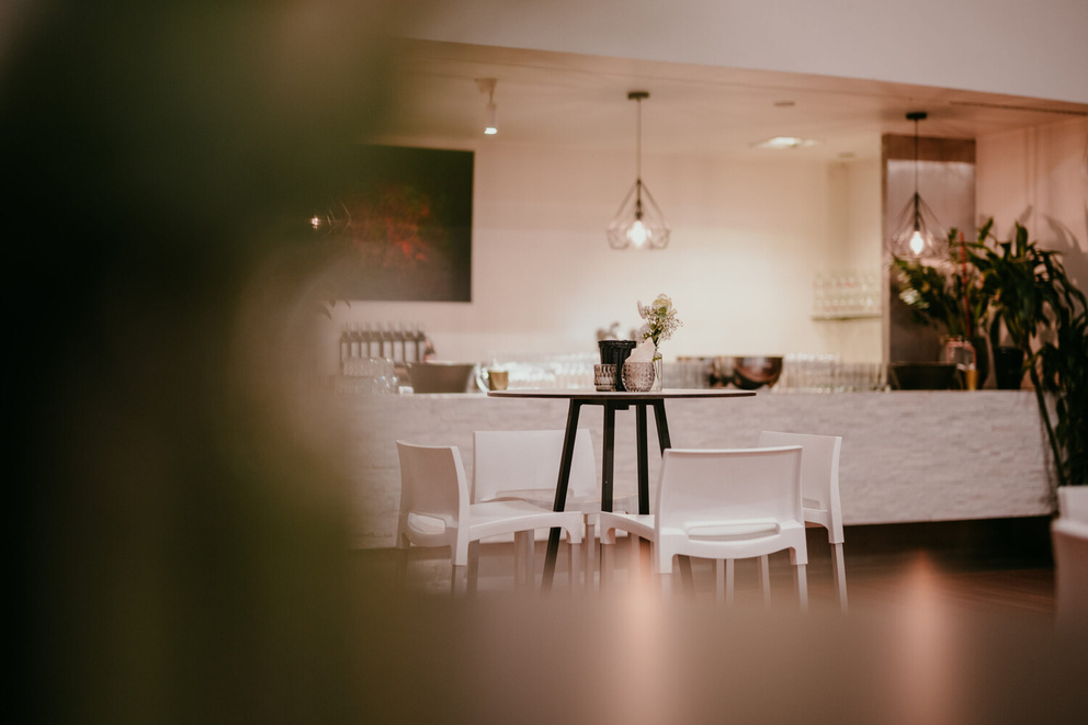 Moderne inrichting van een restaurant met een bar, witte stoelen en groene planten op de achtergrond.