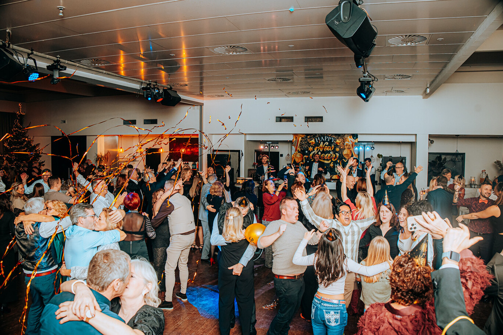 Feestelijke menigte danst in een levendige zaal met ballonnen en confetti. Gelach en vreugde vullen de ruimte.
