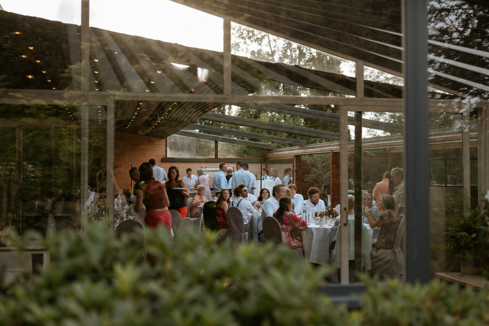 Een elegante zaal vol gasten die aan tafels dineren, omringd door planten en een glazen wand.