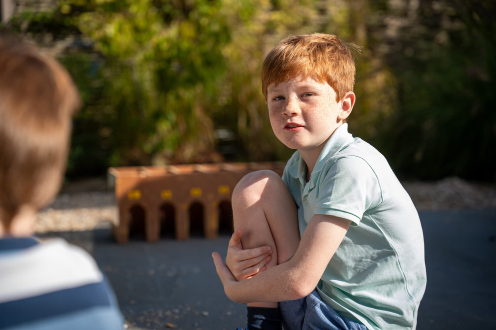 Een jongen met rood haar en sproeten zit op de grond en kijkt ge&iuml;nteresseerd naar iemand naast hem.