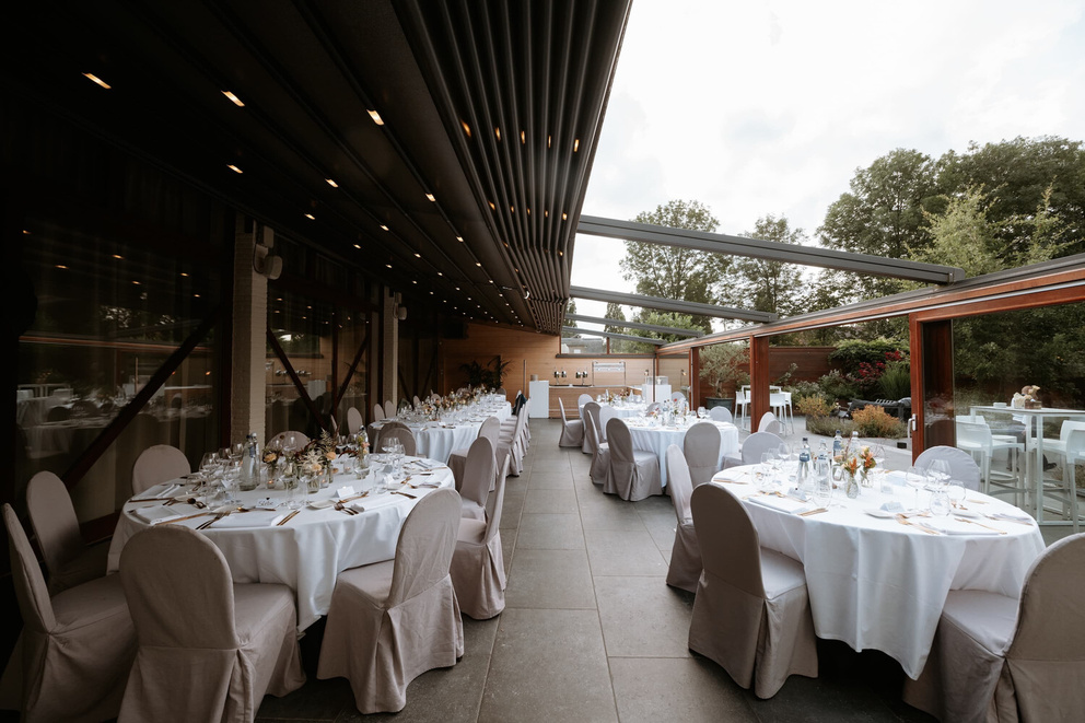 Elegant ingericht restaurant met ronde tafels, witte tafelkleden en een glazen wand die uitzicht biedt op de tuin.