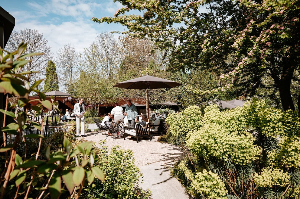 Gezellig terras in de natuur met zonnige parasols, gasten en groene planten in een sfeervolle tuin.