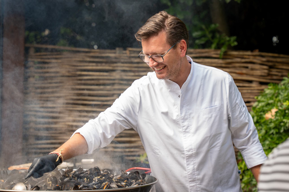 Een chef-kok bereidt mosselen in een grote pan, met glimlach en rokende stoom op de achtergrond.