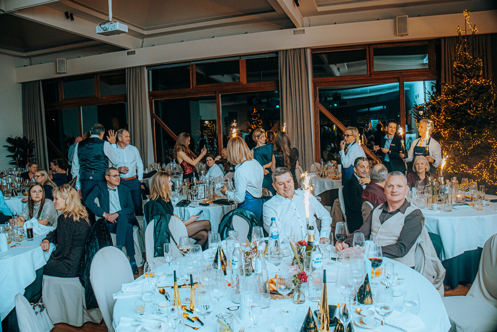 Drukke feestlocatie met gasten aan tafels, versierd voor een feestelijke gelegenheid. Feeststemming met kerstboom op achtergrond.
