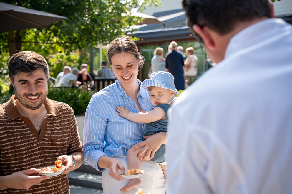 Een man en een vrouw met een baby genieten van eten op een druk terras in de zon.