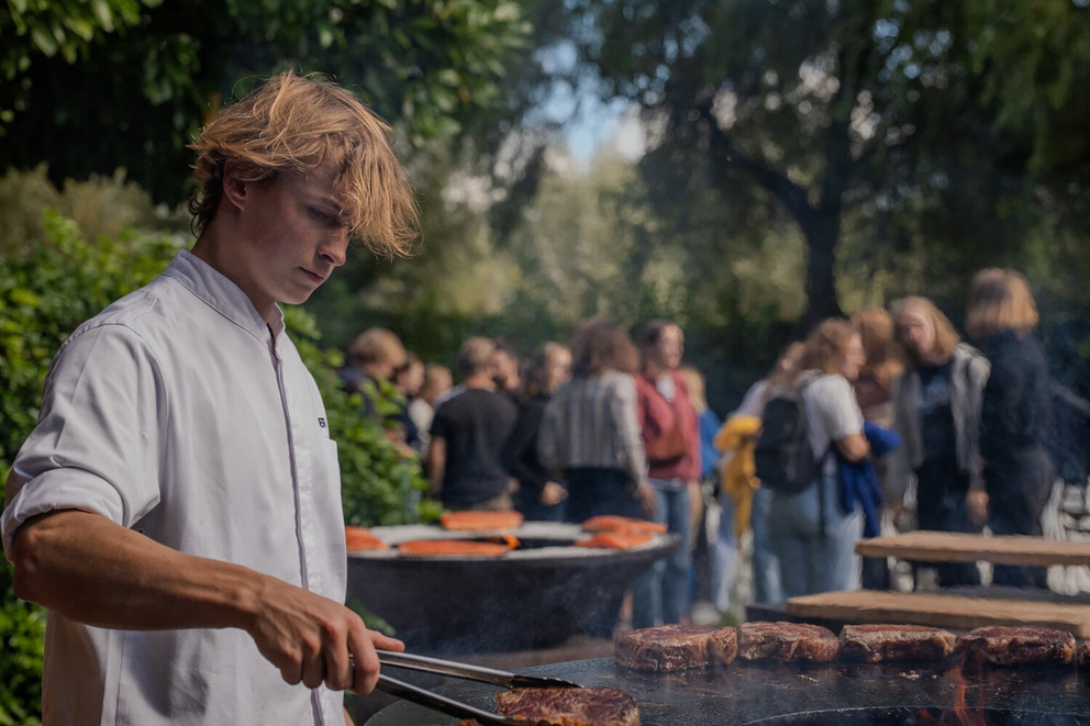 Een jonge chef-kok bereidt vlees op een barbecue, terwijl een groep mensen in de achtergrond geniet van het evenement.