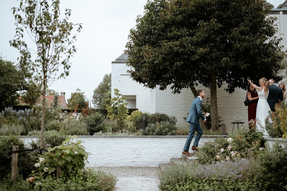Een man in een blauw pak loopt een trap op terwijl een vrouw in een witte jurk enthousiast naar hem lacht in een tuin.